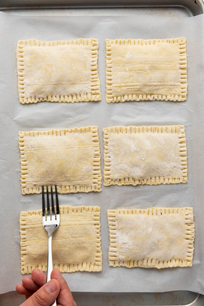 Hand pressing fork along the edges of puff pastry rectangles to seal upside down apple tarts