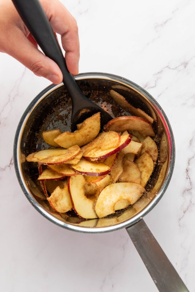 Hand stirring apple slices in a saucepan as they caramelize with sugar and cinnamon