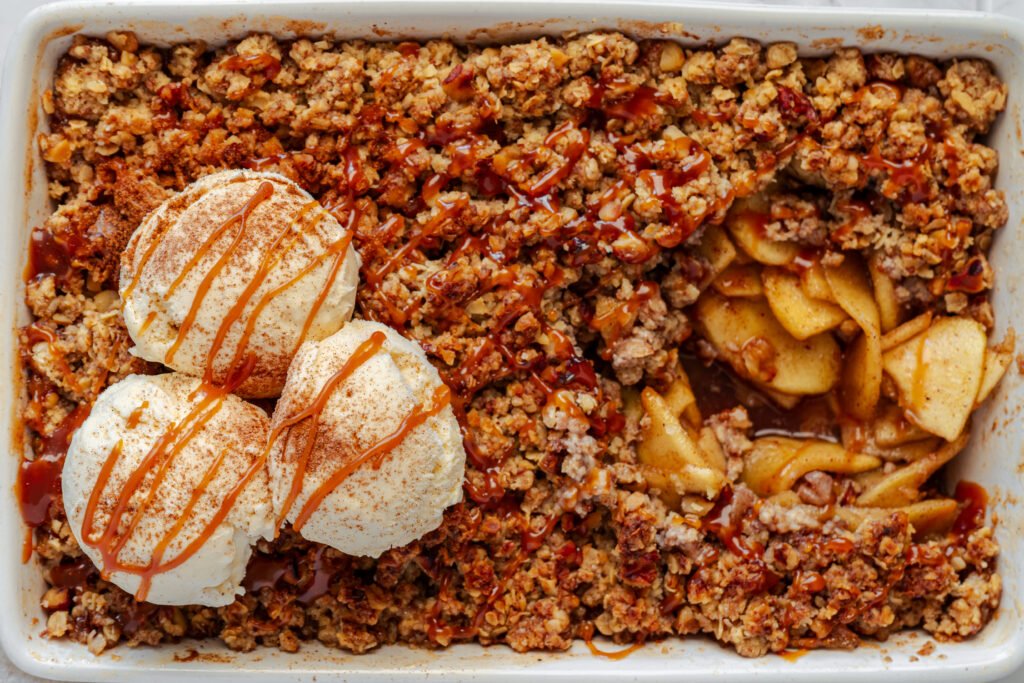 Close-up of apple crisp in a baking dish with scoops of vanilla ice cream and caramel drizzle on top