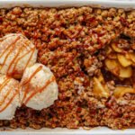 Close-up of apple crisp in a baking dish with scoops of vanilla ice cream and caramel drizzle on top