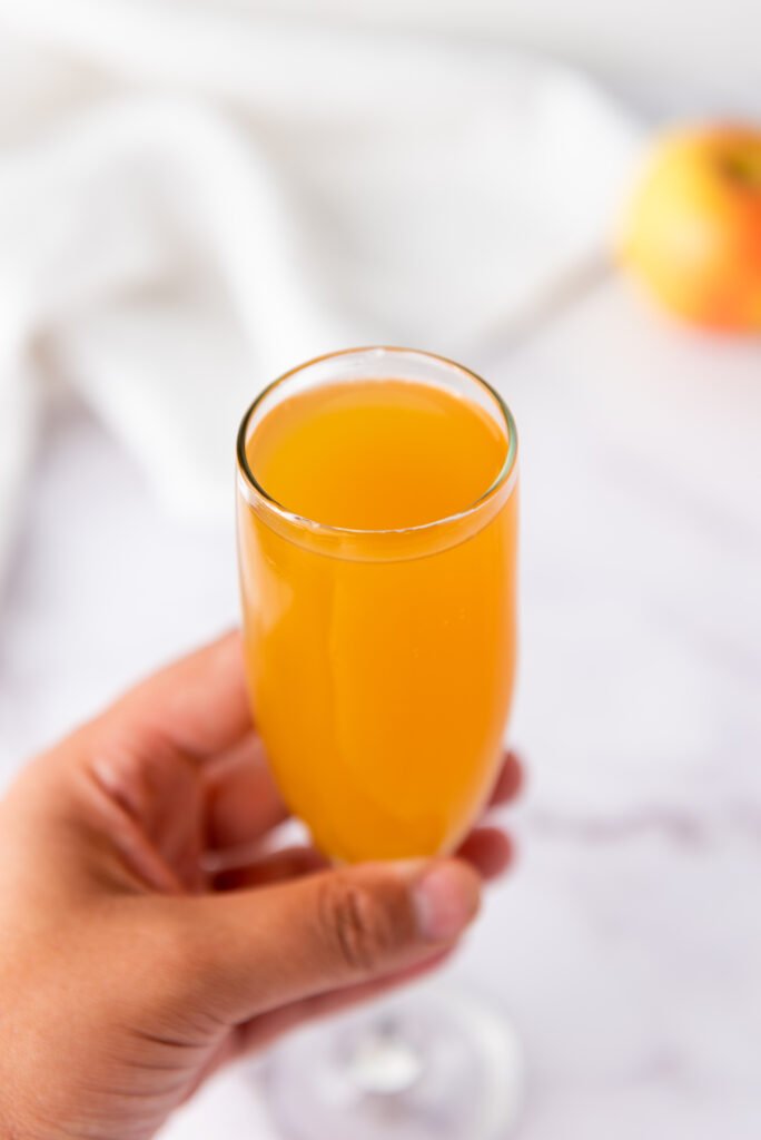 A hand holding a champagne flute filled with golden apple cider mimosa, with a blurred apple in the background