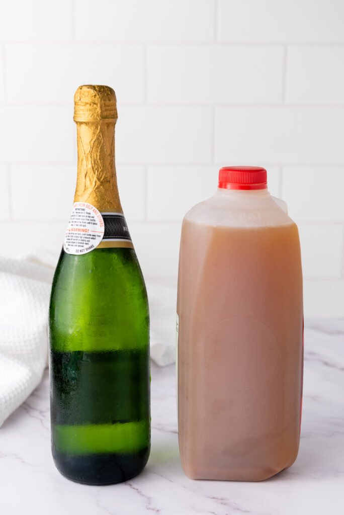A chilled bottle of champagne next to a plastic jug of apple cider, two ingredients for making apple cider mimosas