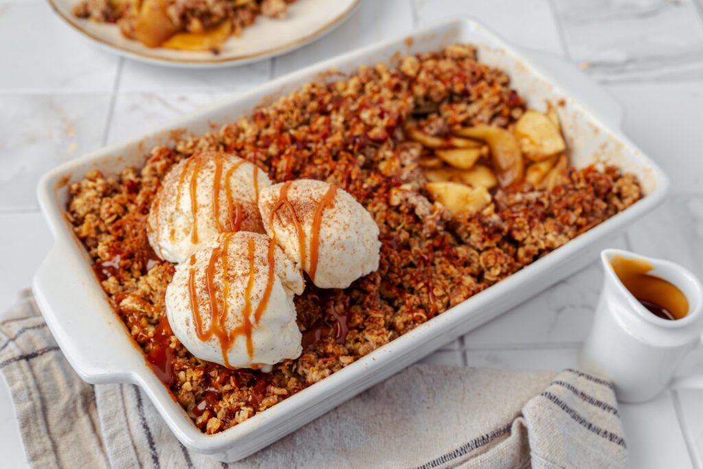 Baked apple crisp in a white dish topped with scoops of vanilla ice cream and drizzled with caramel sauce