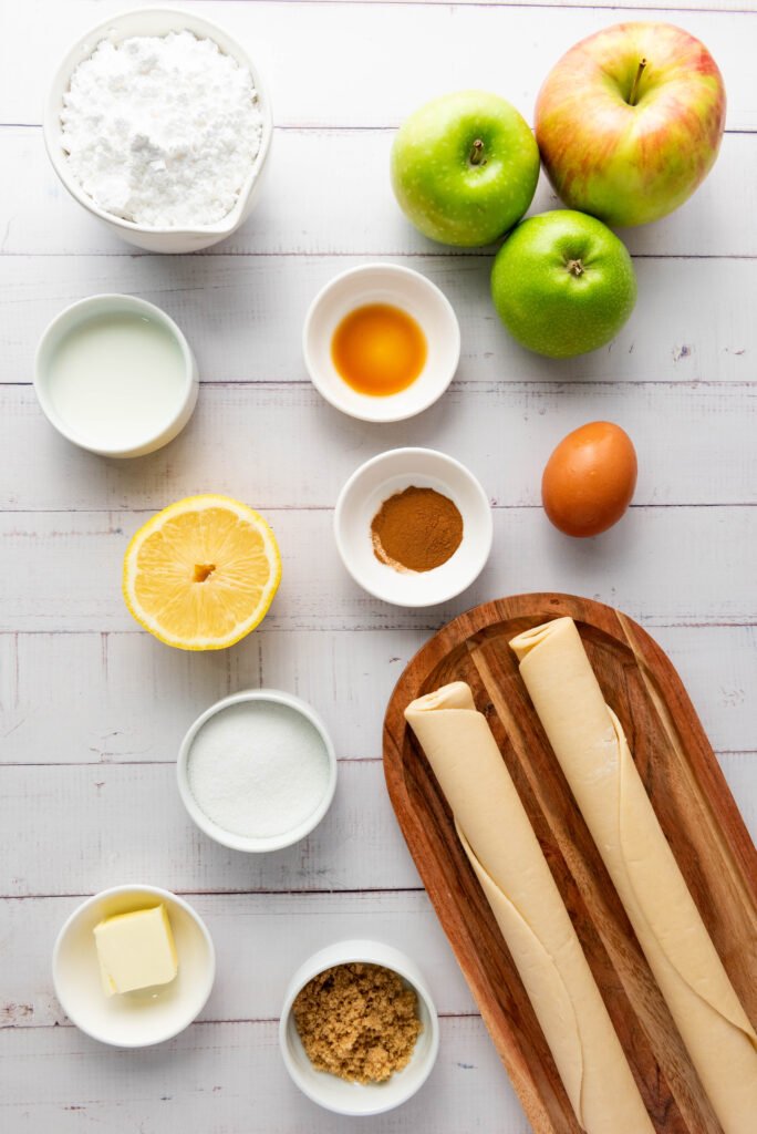 Ingredients for apple hand pies, including apples, lemon, egg, butter, sugars, cinnamon, vanilla, milk, powdered sugar, and rolled pie crusts on a wooden tray