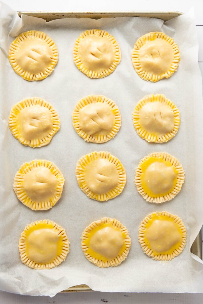 Unbaked apple hand pies brushed with egg wash and arranged neatly on a parchment-lined baking tray