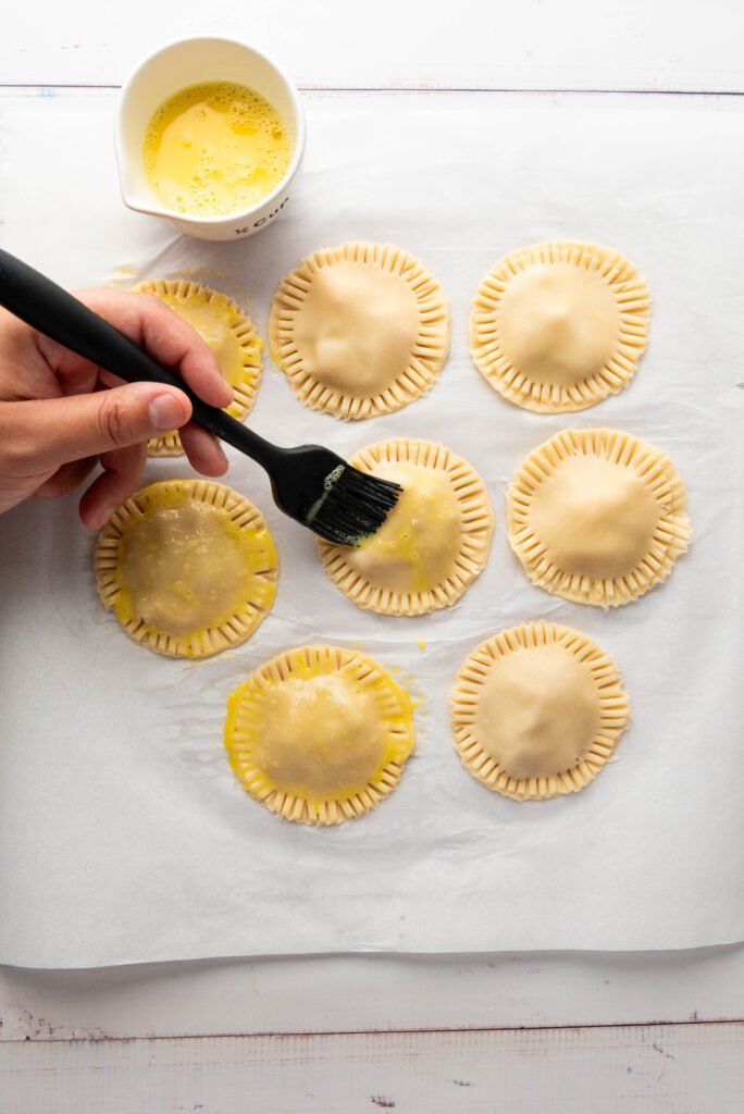 Hand brushing egg wash over unbaked apple hand pies with a pastry brush