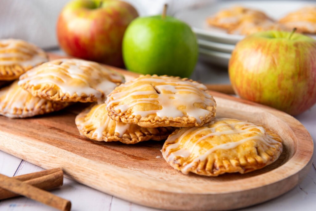 apple hand pies drizzled with vanilla glaze, stacked on a wooden tray with fresh apples and cinnamon sticks in the background