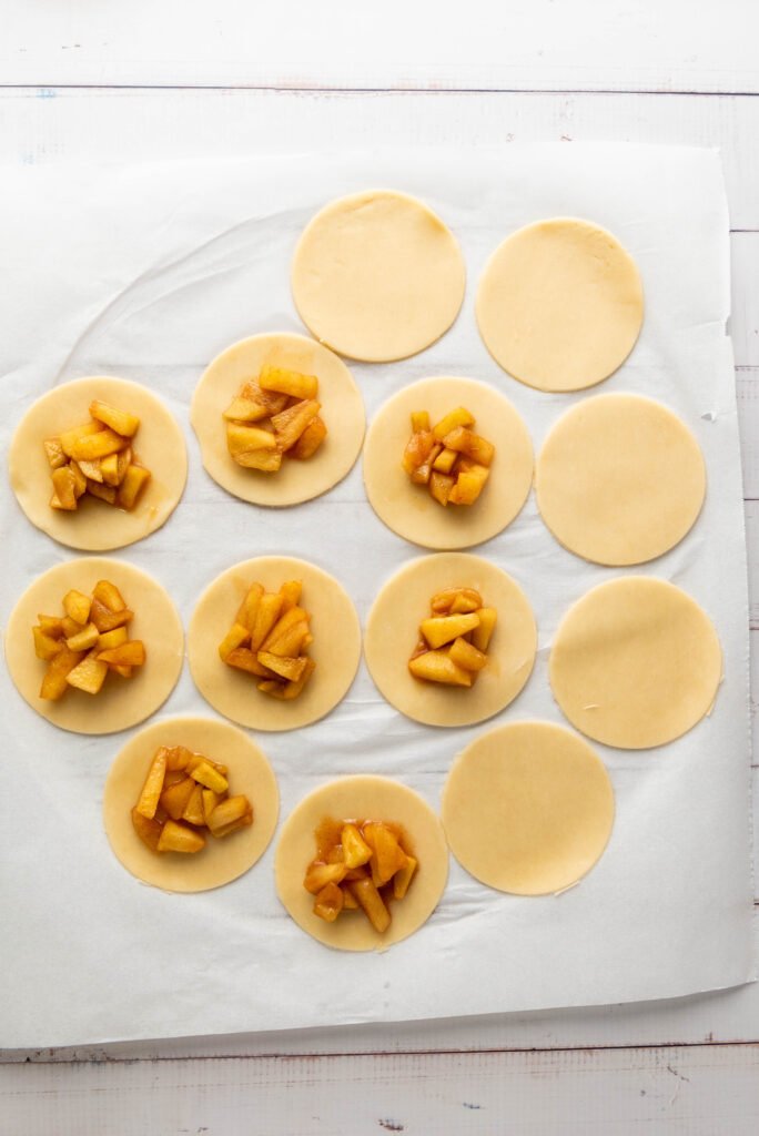 Unbaked apple hand pies with spiced apple filling placed in the center of round pie dough circles on parchment paper