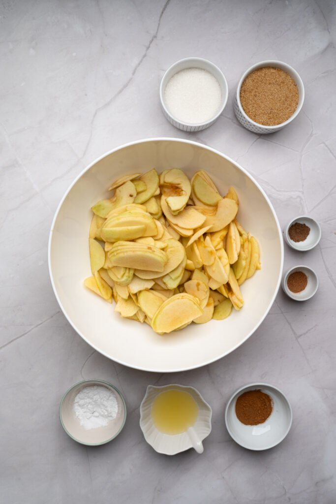Sliced apples in a large bowl surrounded by sugar, spices, cornstarch, and lemon juice for apple crisp filling