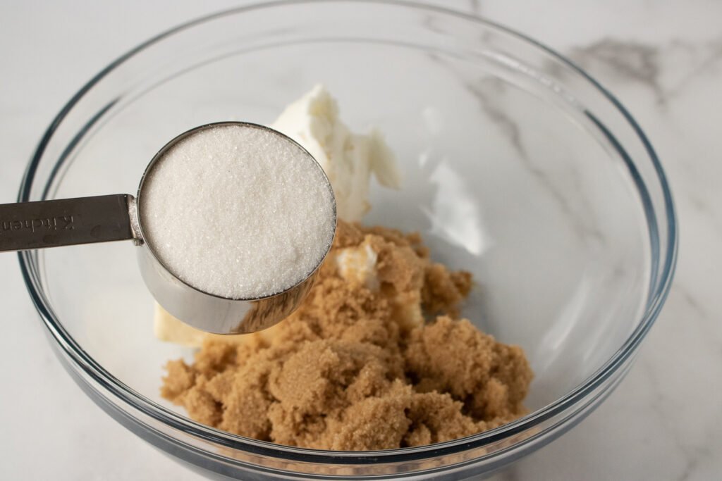 A measuring cup of white sugar being held over a bowl with brown sugar and shortening