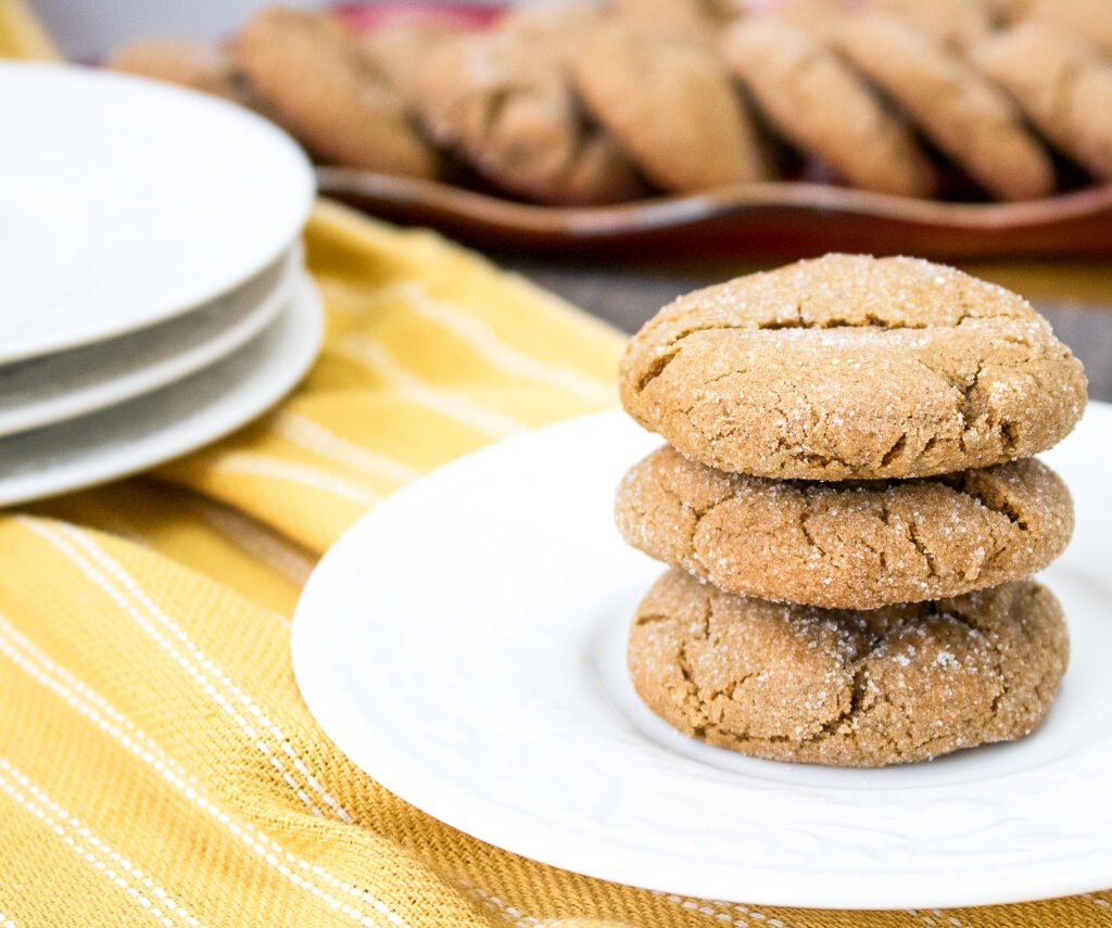 Three chewy gingersnap cookies stacked on a white plate, with more cookies in the background and a yellow striped cloth underneath
