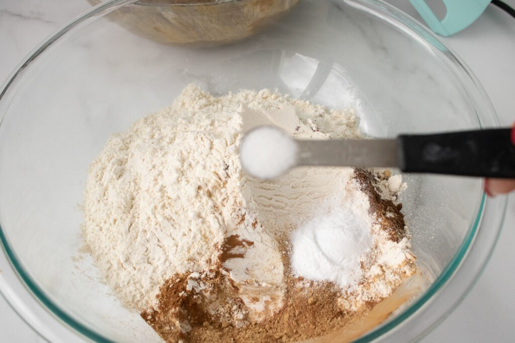 Dry ingredients like flour, baking soda, salt, and warm spices being measured and added to a glass mixing bowl
