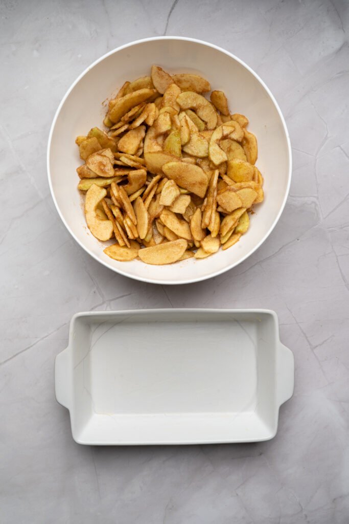 Cinnamon-coated apple slices in a mixing bowl with an empty baking dish ready for filling