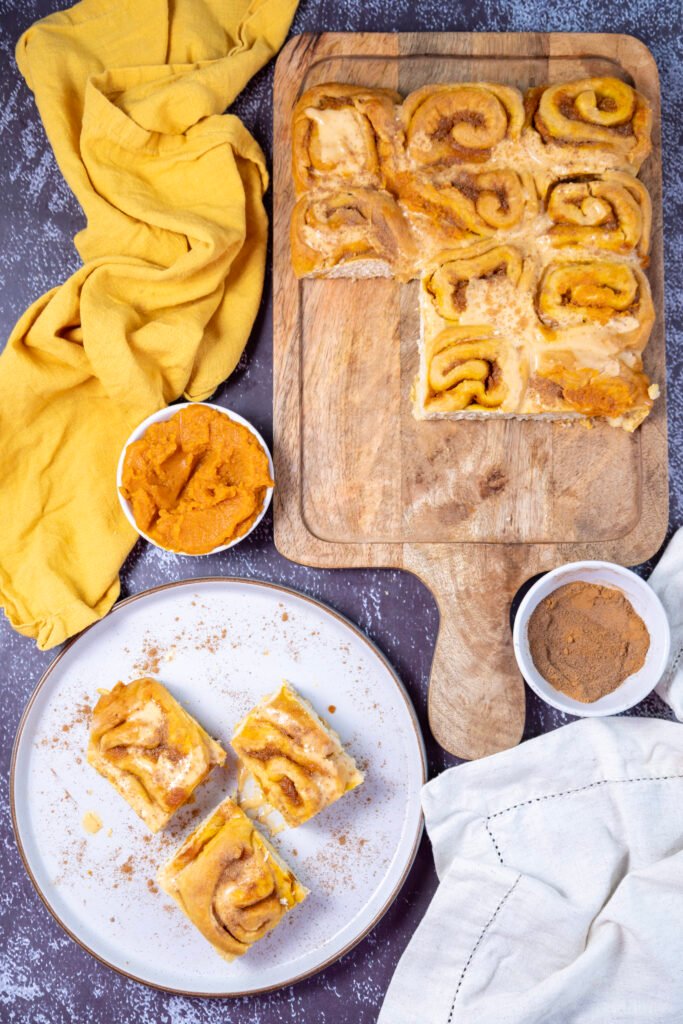 Overhead view of pumpkin cinnamon rolls on a wooden board and plate, with bowls of pumpkin purée and cinnamon nearby