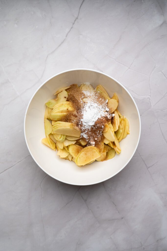 Bowl of sliced apples topped with sugar, cornstarch, and spices before mixing for apple crisp filling