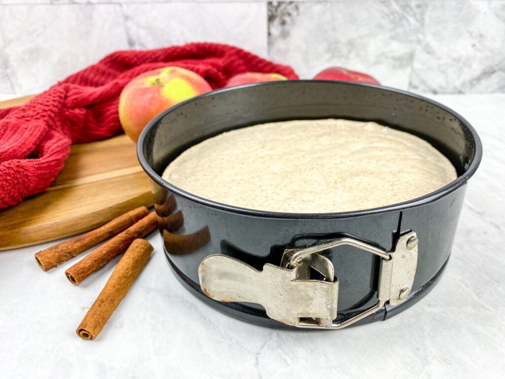 Unbaked apple cinnamon cheesecake batter in a springform pan, surrounded by cinnamon sticks, apples, and a red kitchen towel