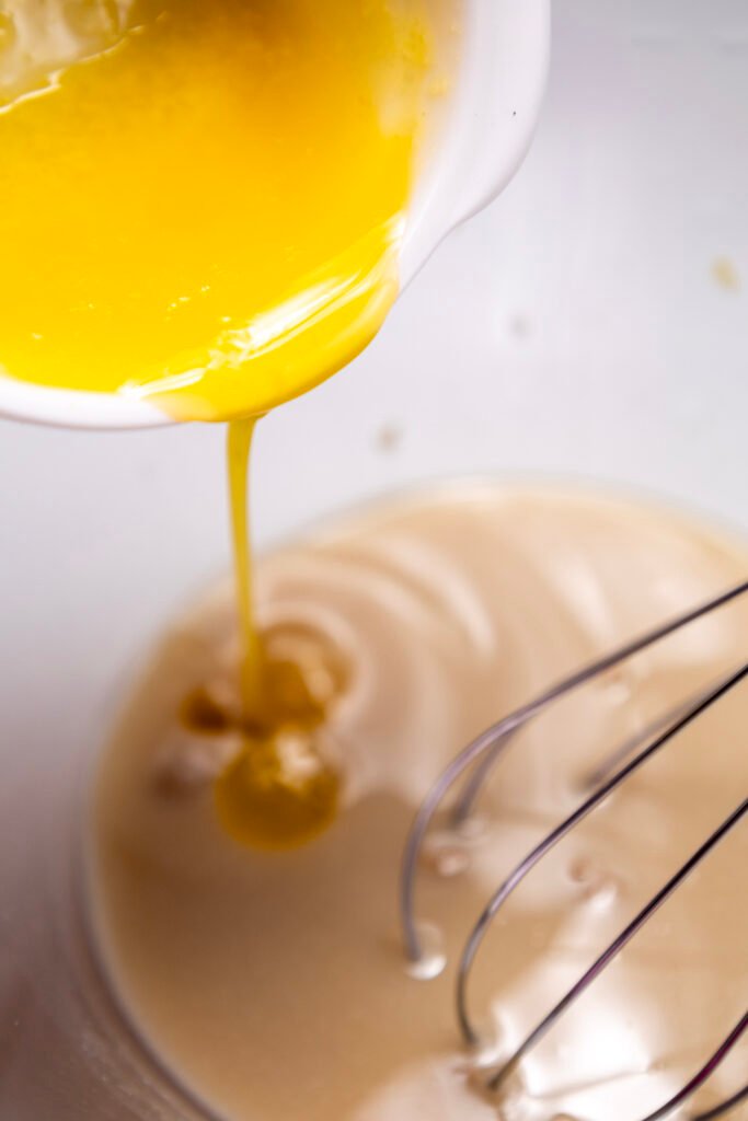 Close-up of melted butter being poured into a bowl of proofed yeast mixture, with a whisk ready for mixing
