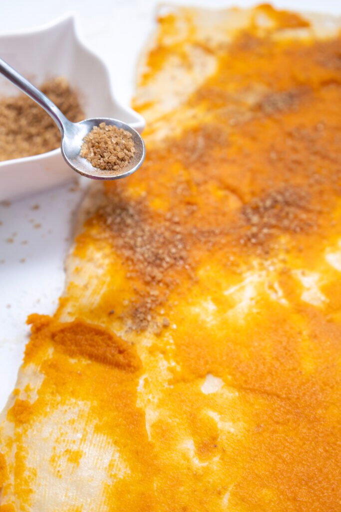 A spoonful of spiced sugar mixture being sprinkled over pumpkin-covered dough