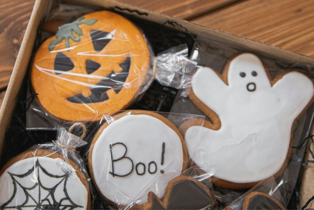 Box of decorated Halloween cookies shaped like a pumpkin, ghost, spiderweb, and a round cookie with “Boo!” written in icing
