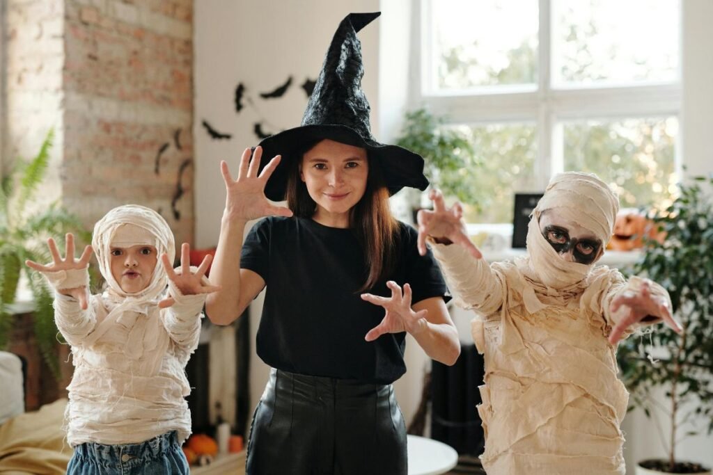 Woman in a witch hat posing with two children dressed as mummies, all making spooky hand gestures during a Halloween party at home