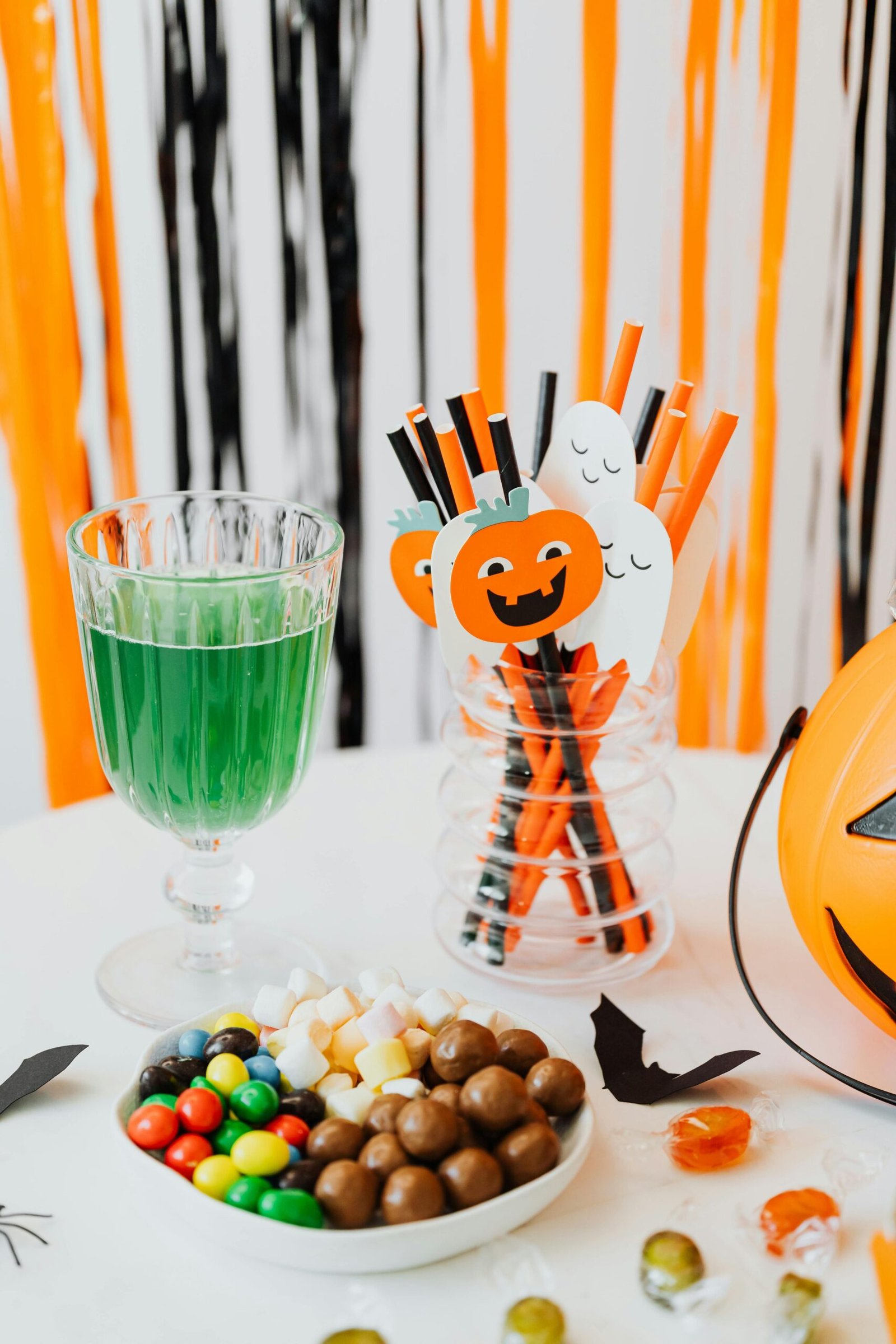 Halloween table with green drink, candy bowl, and festive pumpkin and ghost straws against orange and black decor