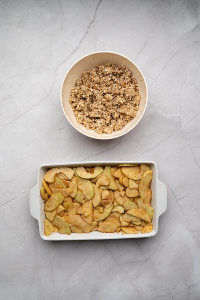 Bowl of oat topping mixture next to a baking dish filled with apple crisp filling, ready to assemble