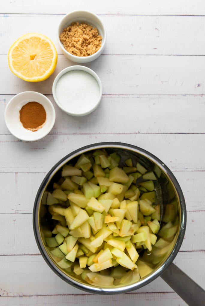 Chopped apples in a saucepan with small bowls of sugar, brown sugar, cinnamon, and half a lemon on the side