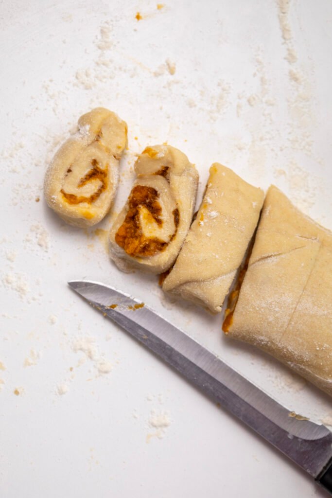 Pumpkin cinnamon roll dough sliced into spirals on a floured surface, with a knife nearby