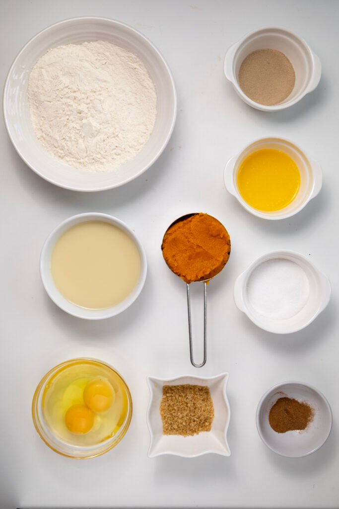 Flat lay of pumpkin cinnamon roll ingredients, including flour, yeast, pumpkin purée, eggs, milk, sugars, melted butter, and spices