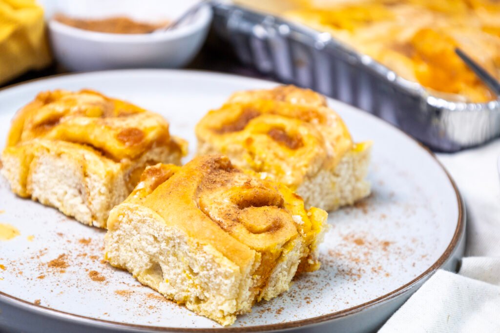 Freshly baked pumpkin cinnamon rolls on a plate, dusted with cinnamon, with more rolls in a foil tray in the background