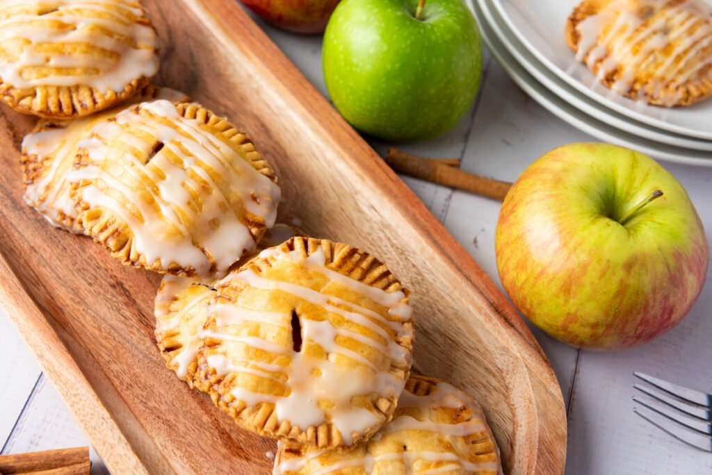 apple hand pies drizzled with vanilla glaze, stacked on a wooden tray with fresh apples and cinnamon sticks in the background