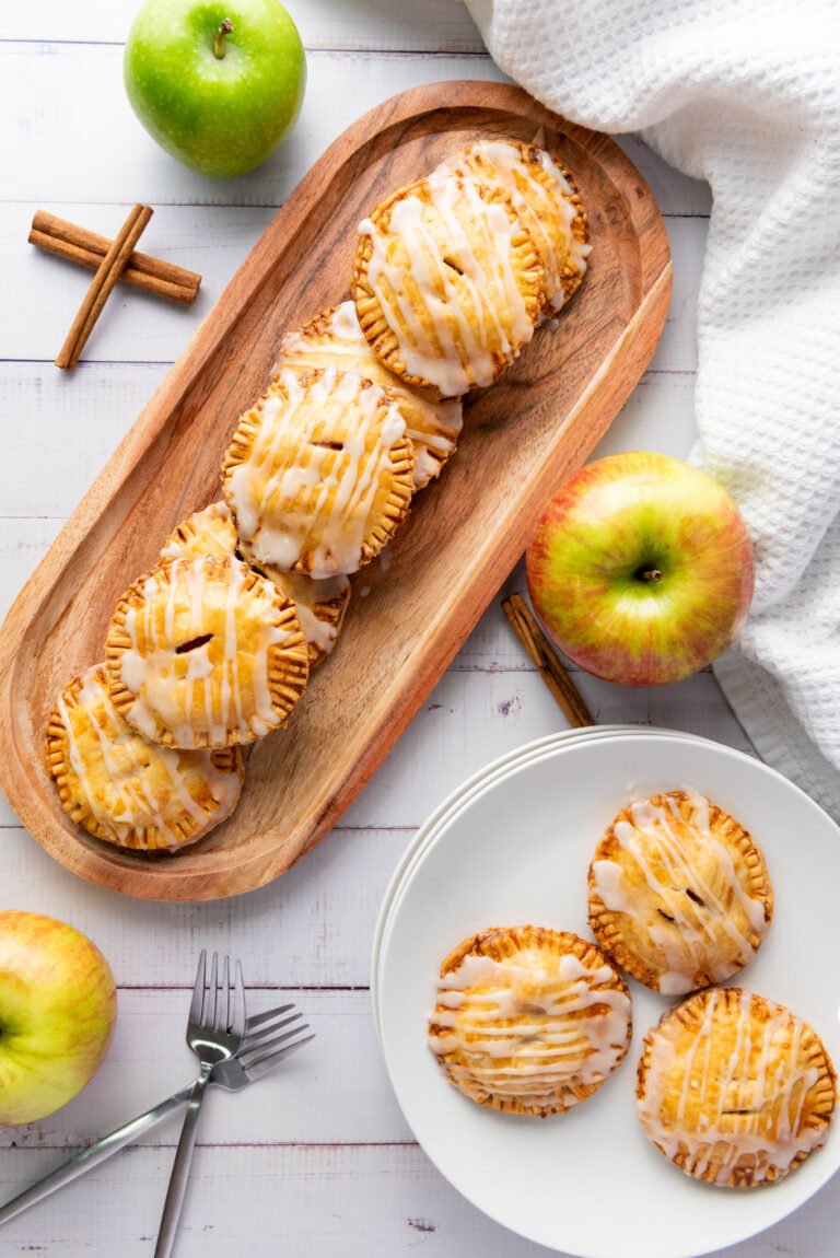 Overhead view of glazed apple hand pies on a wooden tray and white plate, surrounded by fresh apples, cinnamon sticks, and forks
