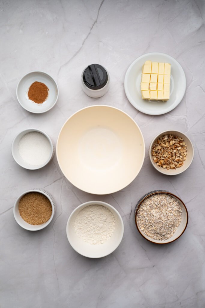 Apple crisp topping ingredients including oats, flour, sugars, butter, cinnamon, salt, and chopped nuts arranged around a mixing bowl