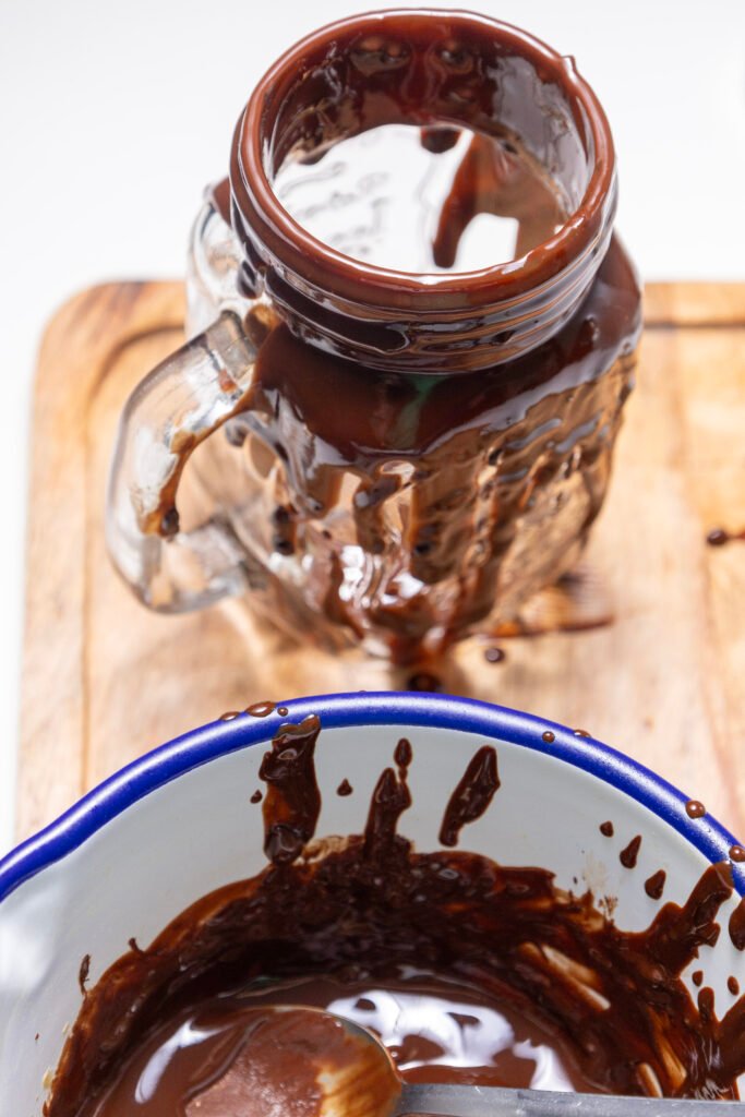 A glass mug covered in melted chocolate, sitting on a wooden board with a bowl of melted chocolate and spoon in the foreground