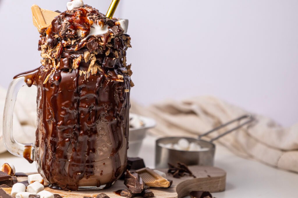 chocolate freakshake in a glass mug, dripping with melted chocolate and topped with whipped cream, marshmallows, chocolate chunks, and caramel pieces