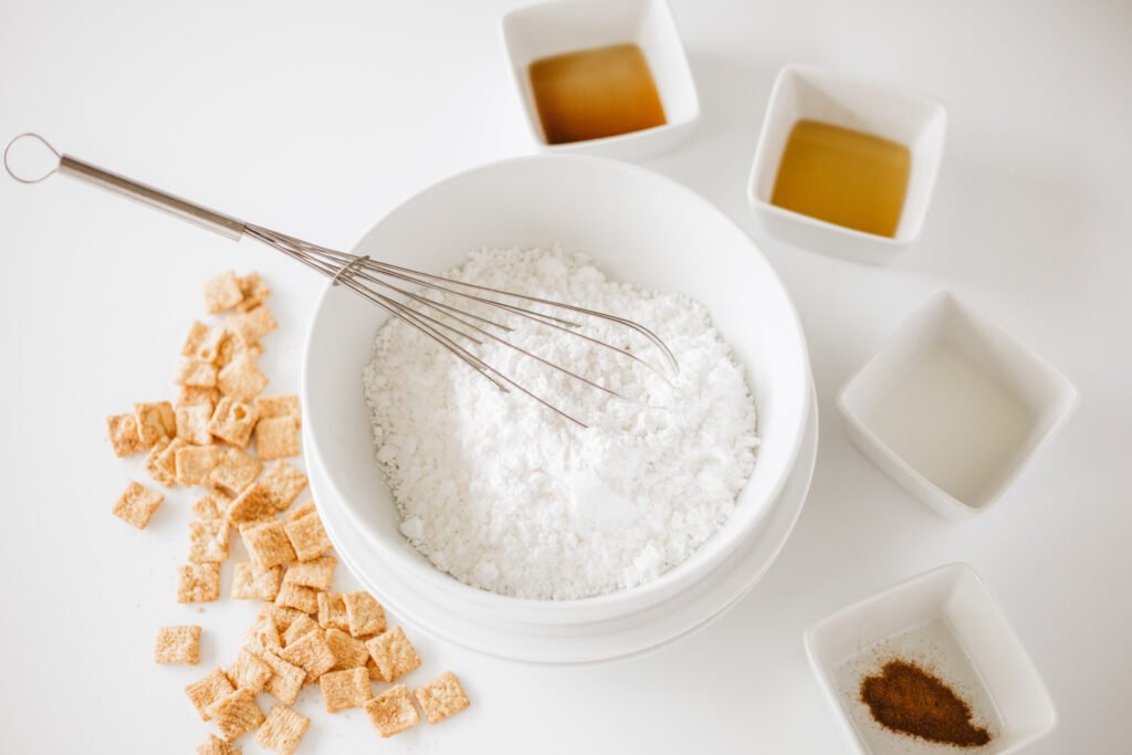 White mixing bowl with powdered sugar and a whisk, surrounded by small bowls of maple syrup