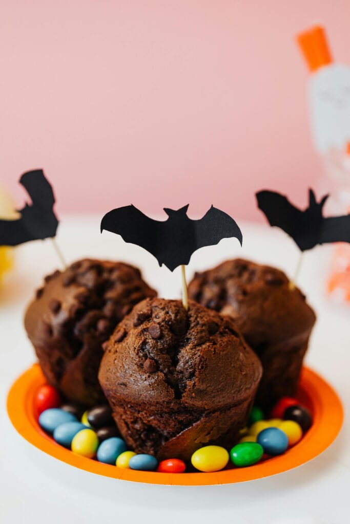 Chocolate muffins decorated with black paper bat toppers, served on an orange plate surrounded by colorful candy-coated chocolates