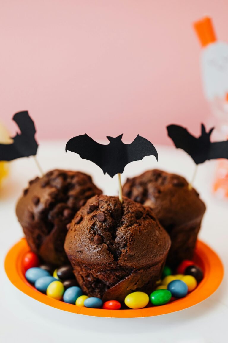 Chocolate muffins decorated with black paper bat toppers, served on an orange plate surrounded by colorful candy-coated chocolates
