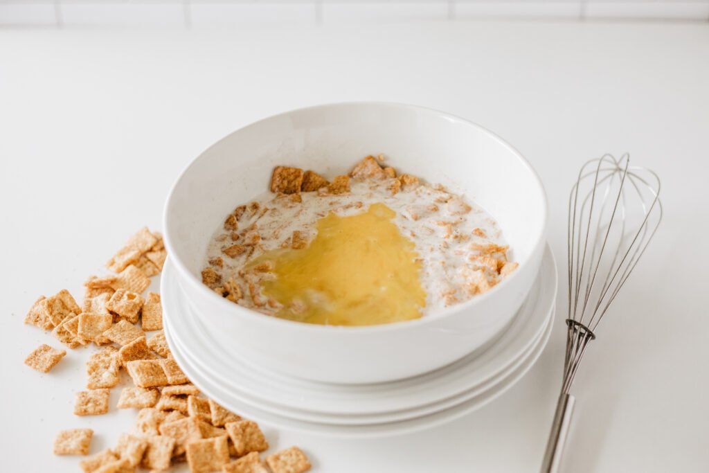 White mixing bowl with crushed Cinnamon Toast Crunch cereal and buttermilk, topped with a cracked egg