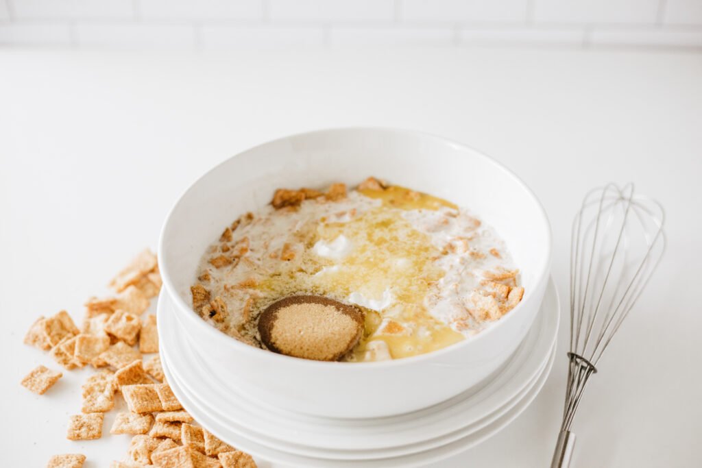 White mixing bowl with cereal and buttermilk mixture, now combined with egg, melted butter, Greek yogurt, and brown sugar
