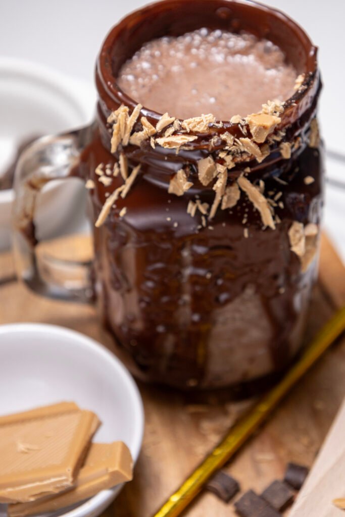 A chocolate freakshake in a glass mug coated with melted chocolate, garnished with blonde chocolate shavings around the rim
