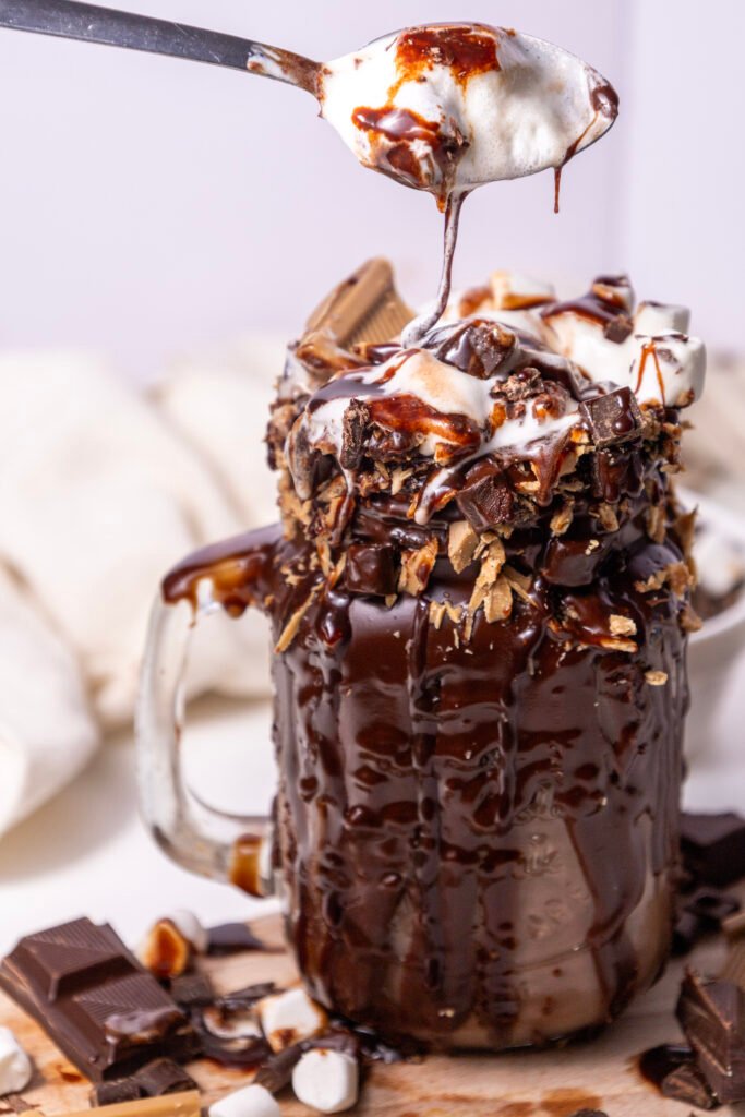 chocolate freakshake in a glass mug, dripping with melted chocolate and topped with whipped cream, marshmallows, chocolate chunks, and caramel pieces