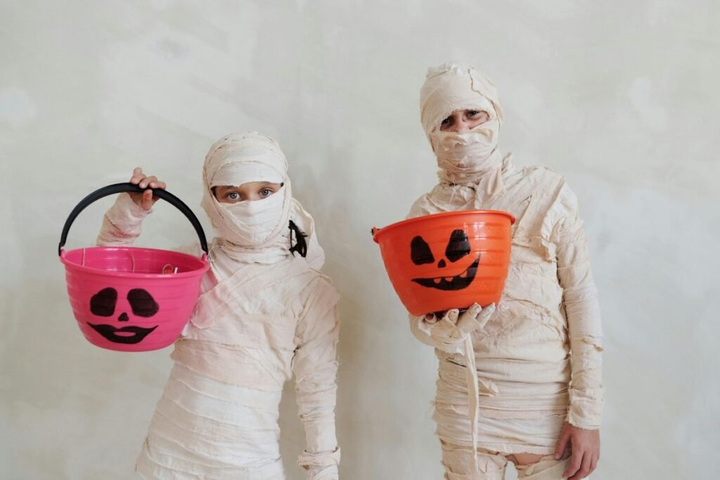 Two children dressed in mummy costumes hold Halloween buckets, one pink and one orange with jack-o’-lantern faces
