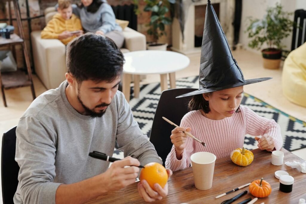 A man and a young girl decorate small pumpkins at a table for Halloween