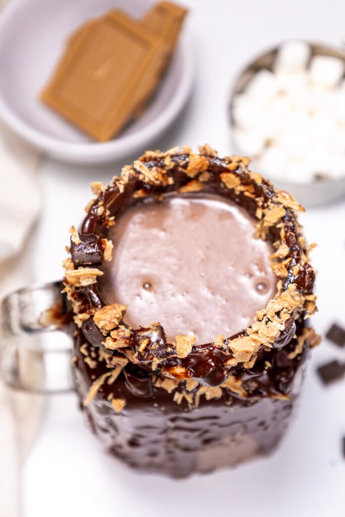Top view of a chocolate freakshake topped with blonde chocolate shavings, with bowls of marshmallows and chocolate pieces in the background