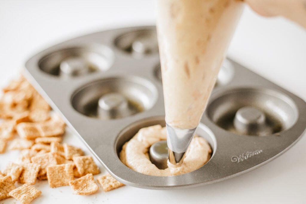 Cinnamon Toast Crunch donut batter being piped into a greased donut pan cavity using a piping bag
